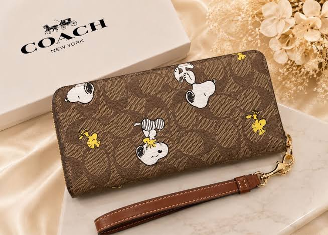 A Coach New York wristlet wallet featuring a brown signature canvas print with Snoopy and Woodstock character illustrations, shown with its white Coach box and a wrist strap on a beige satin background.