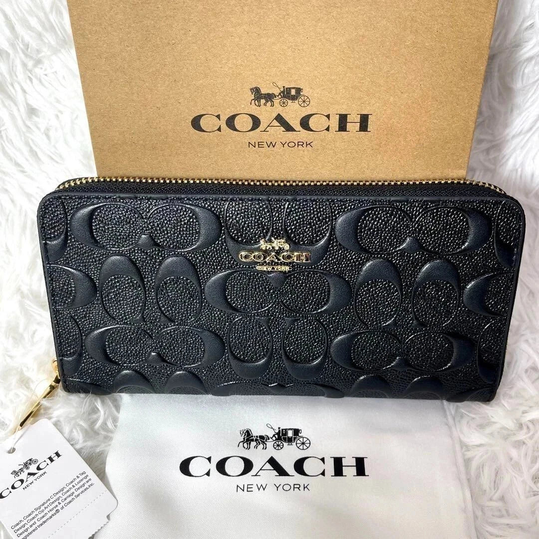 A black Coach zip-around wallet with the signature monogram pattern, displayed in front of a kraft paper Coach box and a white dust bag on a white faux-fur surface.