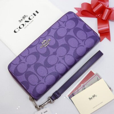A purple Coach zip-around wallet with the signature monogram pattern, a wristlet strap, and a small gold-tone logo clasp, displayed on a white surface with a red ribbon, shopping box, and cards.