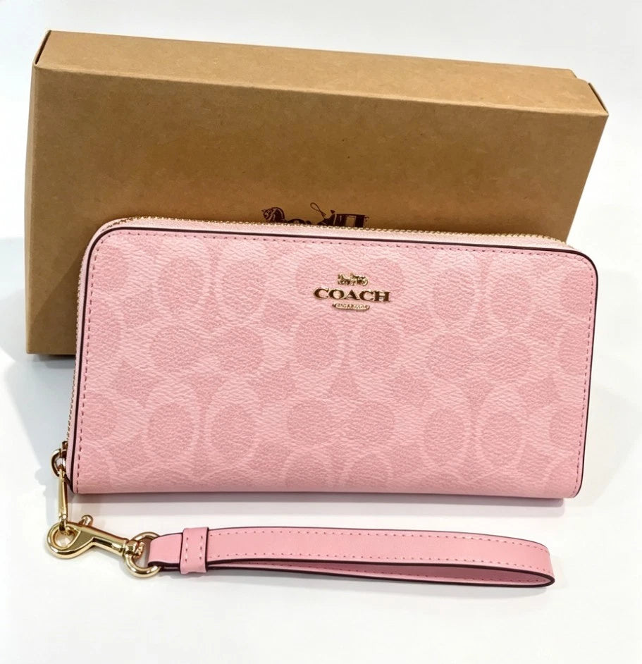 A pink Coach wristlet wallet with the signature monogram pattern and gold-tone logo, shown with its brown cardboard packaging box on a white background.