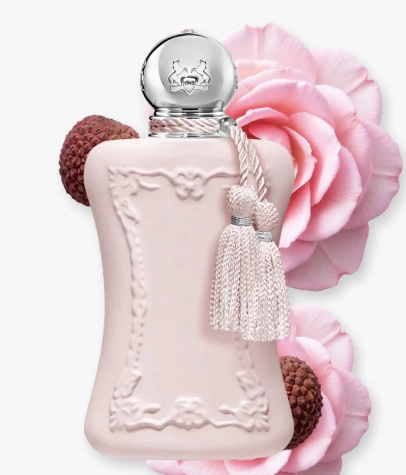 A pale blush pink perfume bottle with an embossed floral border, a silver-toned round cap, and a tassel charm, styled with pink roses and brown textured accents on a light gray background.